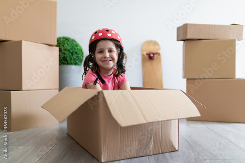 Happy little girl smiling and fastening helmet while sitting in cardboard box and playing during relocation photo