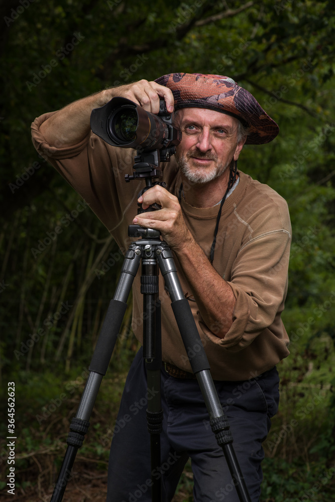 Photographer mature man gray beard floppy hat brown shirt blue pants outside, green wooded area, looking out  from behind DSLR digital camera with zoom telephoto lens on tripod
