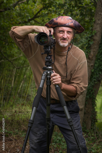 Photographer mature man gray beard floppy hat brown shirt blue pants outside, green wooded area, ready to snap photo from behind DSLR digital camera with zoom telephoto lens on tripod