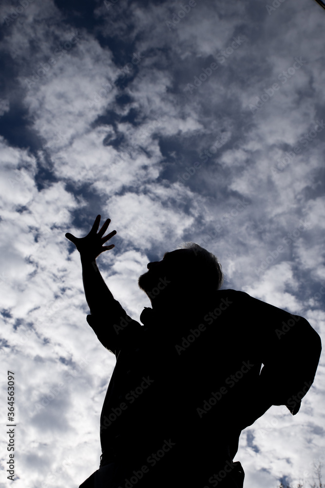 Male silhouette looking up at and one hand extending reaching up stretching trying to touch the blue and white cloud filled sky