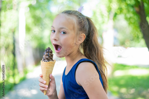 Happy little caucasian girl in blue dress eats chocolate ice cream cone outdoors. An emotional excited child enjoys a cooling gelato on a hot summer day.