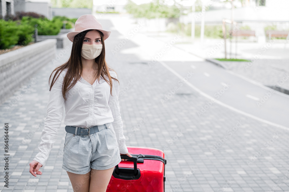 A beautiful young Caucasian woman in a medical mask walks down the street with a red suitcase. A girl with luggage travels alone during the height of the coronavirus epidemic. The empty city. A beautiful young Caucasian woman in a medical mask walks down the street with a red suitcase. A girl with luggage travels alone during the height of the coronavirus epidemic. The empty city.