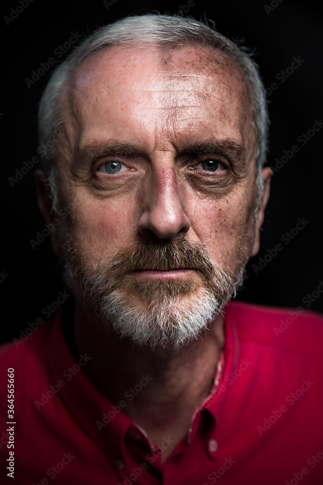 Middle aged male split aging portrait headshot with gray hair and beard red shirt on black background and half face high contrast to simulate aging or weathered look and age spots Middle aged male split aging portrait headshot with gray hair and beard red shirt on black background and half face high contrast to simulate aging or weathered look and age spots