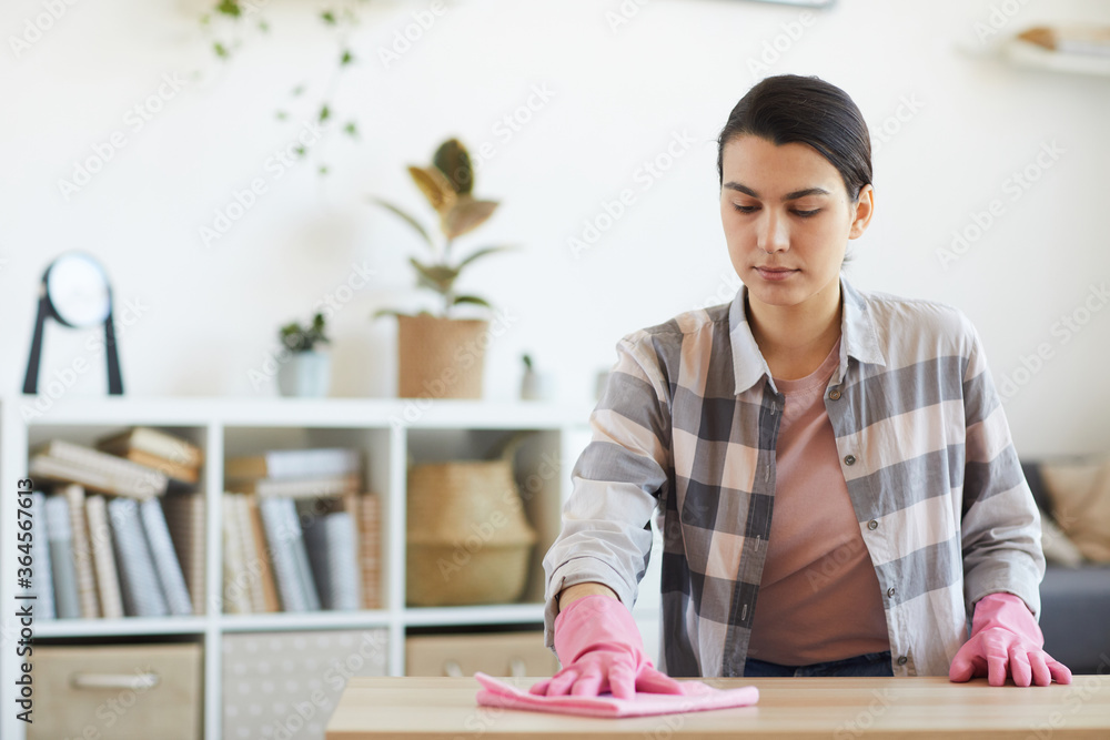 Young housekeeper in rubber gloves wiping dust from the table with rag in the room Young housekeeper in rubber gloves wiping dust from the table with rag in the room