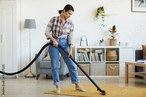 Young woman cleaning the carpet with vacuum cleaner in the room at home