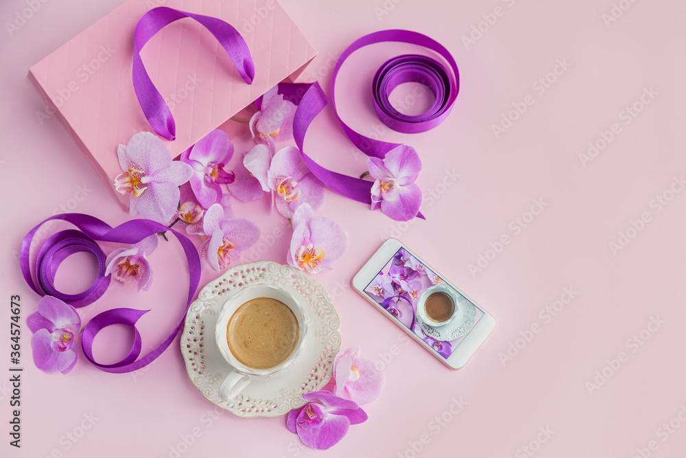 Obraz premium Flower overhead composition on light pink background top view. Cup of coffee, pink gift bag with purple ribbons, phone and pink orchid flowers.