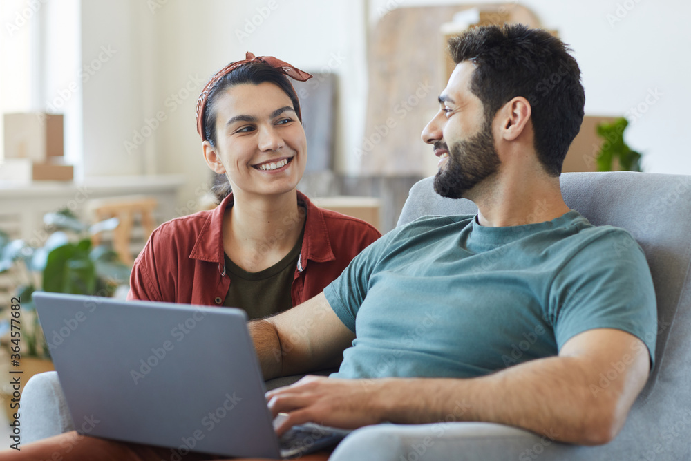 Young happy couple sitting on sofa talking to each other and using laptop while resting at home
