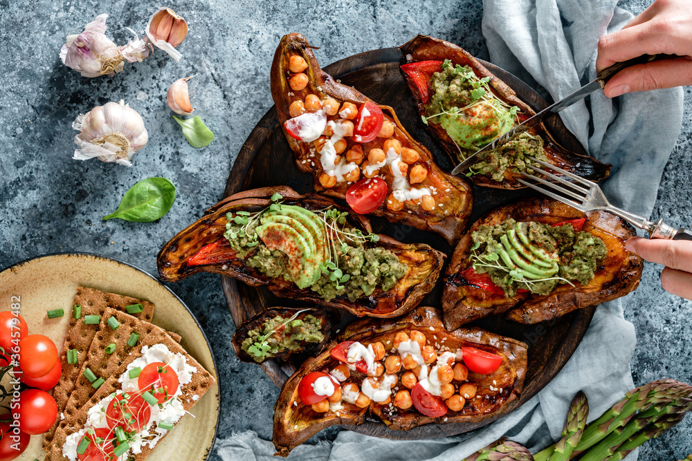 Hands cutting baked sweet potato toasts with roasted chickpeas, tomatoes, avocado, seedlings on wooden board. Healthy vegan food, clean eating, dieting, top view