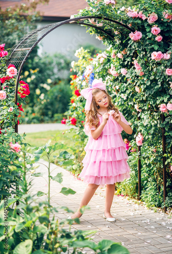 girl in a pink dress with a bow on her head in the rose garden. Girl doll