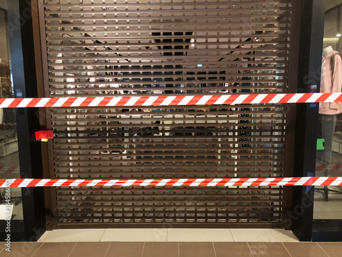 striped boundary tape covers brown shutted iron gates of shop in shopping mall due to restrictions of lockdown. Protection measures against coronavirus pandemic.bankruptcy of stores