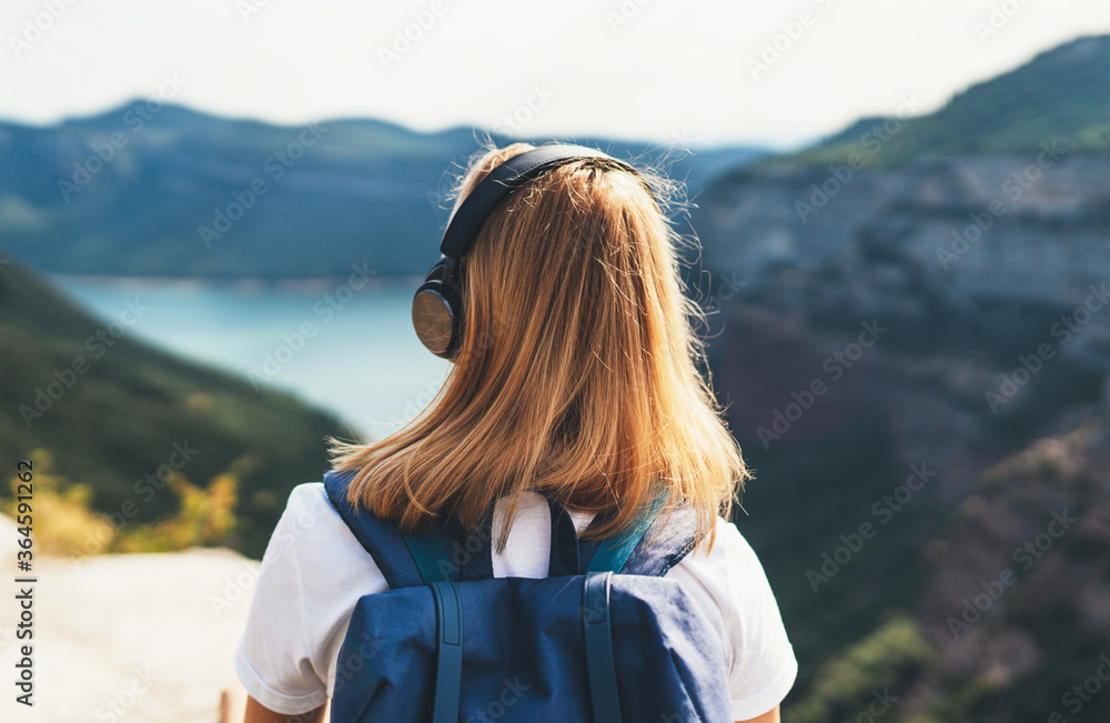 back view girl tourist with light hair dreams listening to music in headphones standing on top of mountains, blonde hipster with backpack walking lake valley and green summer hills, copy space back view girl tourist with light hair dreams listening to music in headphones standing on top of mountains, blonde hipster with backpack walking lake valley and green summer hills, copy space