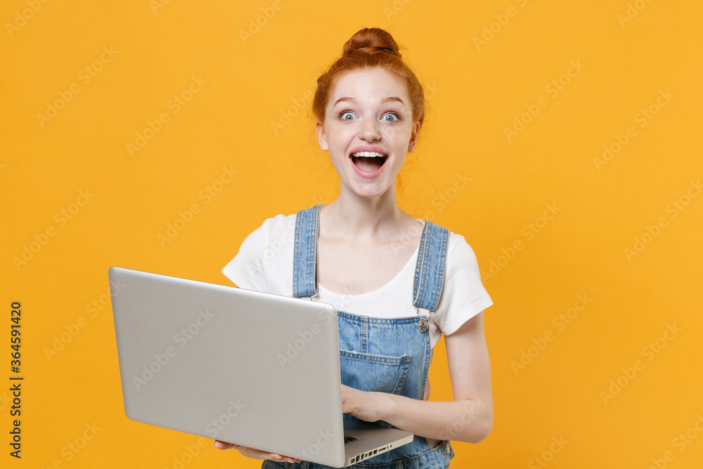 Naklejka premium Surprised young readhead girl in casual denim clothes white t-shirt isolated on yellow background studio portrait. People emotions lifestyle concept. Mock up copy space. Working on laptop pc computer.