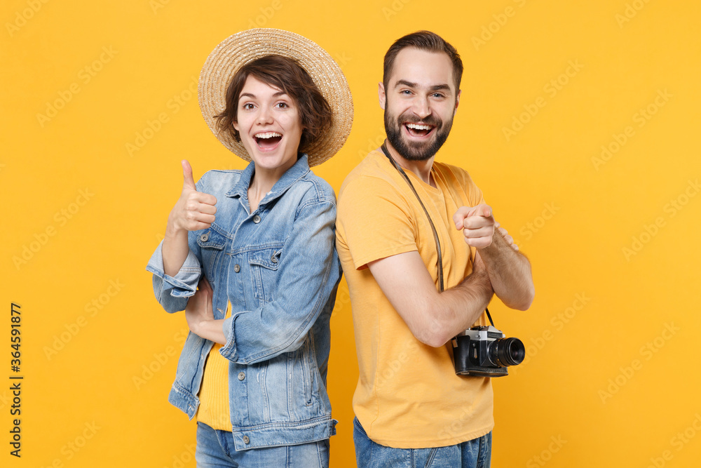 Funny tourists couple friends guy girl with photo camera isolated on yellow background. Passenger traveling abroad on weekends. Air flight journey concept. Pointing finger on camera showing thumb up. Funny tourists couple friends guy girl with photo camera isolated on yellow background. Passenger traveling abroad on weekends. Air flight journey concept. Pointing finger on camera showing thumb up.