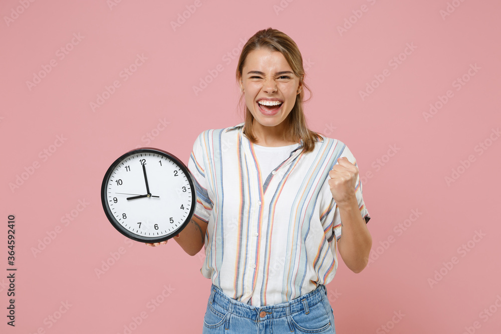 Happy young blonde woman girl in casual striped shirt posing isolated on pastel pink background studio portrait. People emotions lifestyle concept. Mock up copy space. Hold clock doing winner gesture. Happy young blonde woman girl in casual striped shirt posing isolated on pastel pink background studio portrait. People emotions lifestyle concept. Mock up copy space. Hold clock doing winner gesture.