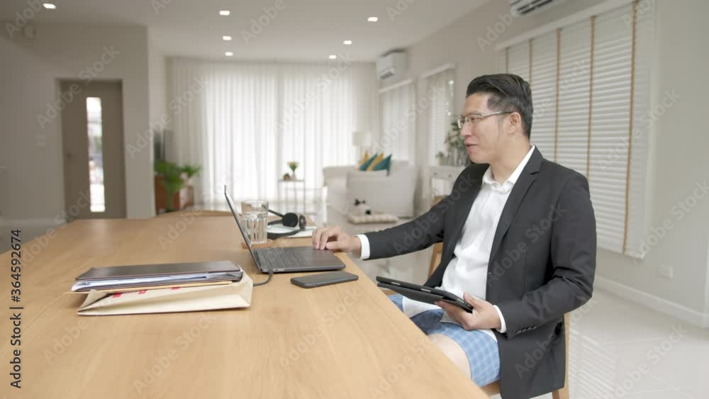 Asian man wearing a suit or business wear on top and sweatpants or boxers on bottom. Businessman video conference using laptop and tablet online meeting.Working from home and Working remotely. Asian man wearing a suit or business wear on top and sweatpants or boxers on bottom. Businessman video conference using laptop and tablet online meeting.Working from home and Working remotely.