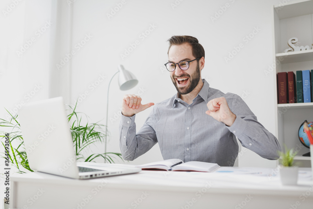 Funny business man in gray shirt glasses sit at desk in light office on white wall background. Achievement business career concept. Work on laptop computer making video call point thumbs on himself.