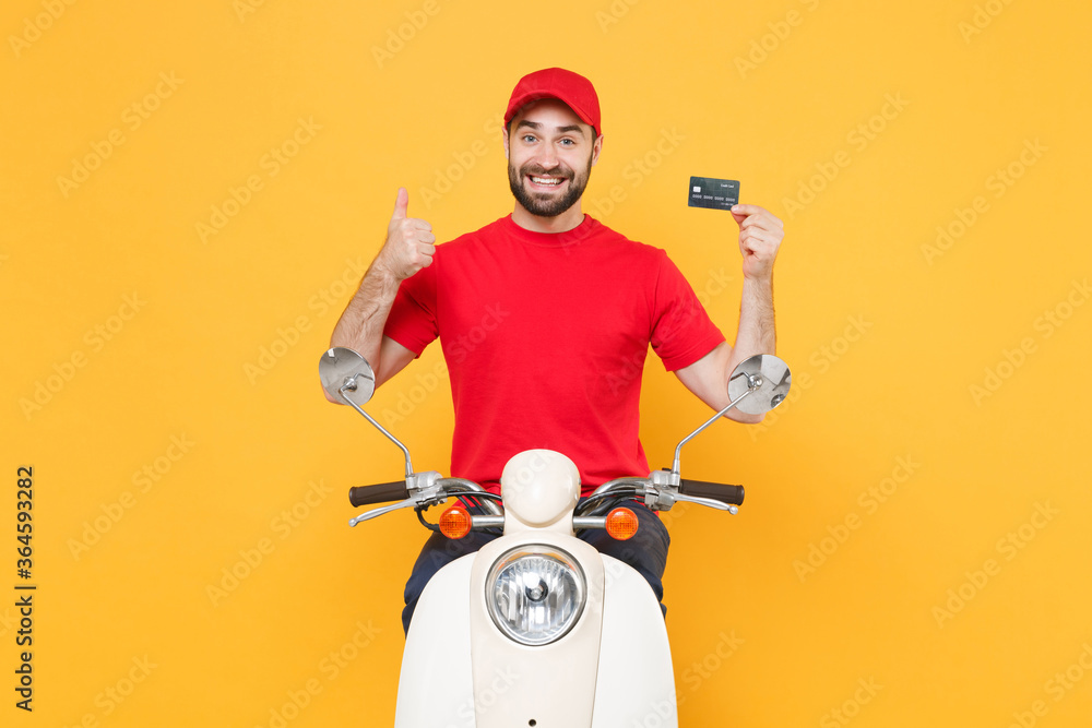 Delivery man in red cap t-shirt uniform driving moped motorbike scooter hold credit card isolated on yellow background studio Guy employee working courier Service quarantine pandemic covid-19 concept