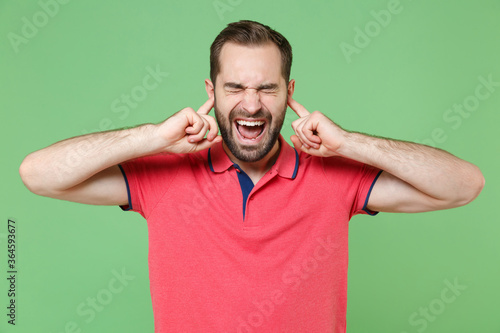 Crazy young bearded man guy in casual red pink t-shirt posing isolated on green background. People lifestyle concept. Mock up copy space. Covering ears with fingers, keeping eyes closed, screaming.