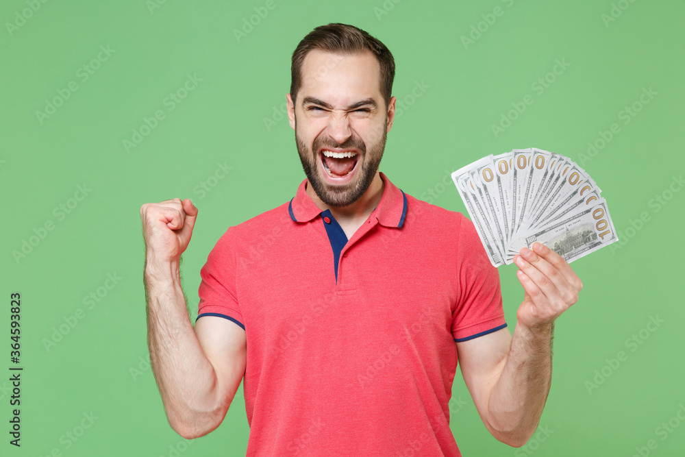 Screaming young bearded man guy in casual red pink t-shirt isolated on green background. People lifestyle concept. Mock up copy space. Hold fan of cash money in dollar banknotes, doing winner gesture. Screaming young bearded man guy in casual red pink t-shirt isolated on green background. People lifestyle concept. Mock up copy space. Hold fan of cash money in dollar banknotes, doing winner gesture.