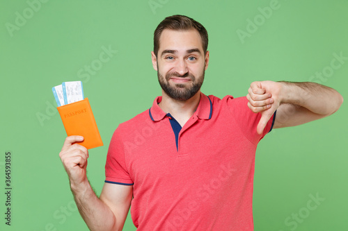 Displeased young guy in red pink t-shirt isolated on green background. Passenger traveling abroad to travel on weekends getaway. Air flight journey concept. Hold passport tickets showing thumb down.