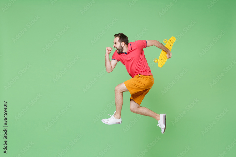 full length Crazy young man guy in casual red pink t-shirt posing isolated on green background studio portrait. People lifestyle concept. Mock up copy space Jumping like running hold yellow skateboard full length Crazy young man guy in casual red pink t-shirt posing isolated on green background studio portrait. People lifestyle concept. Mock up copy space Jumping like running hold yellow skateboard