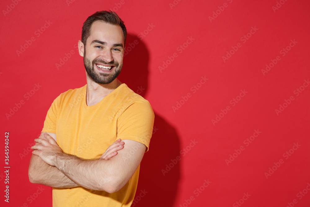 Cheerful young bearded man guy in casual yellow t-shirt posing isolated on red wall background studio portrait. People sincere emotions lifestyle concept. Mock up copy space. Holding hands crossed. Cheerful young bearded man guy in casual yellow t-shirt posing isolated on red wall background studio portrait. People sincere emotions lifestyle concept. Mock up copy space. Holding hands crossed.