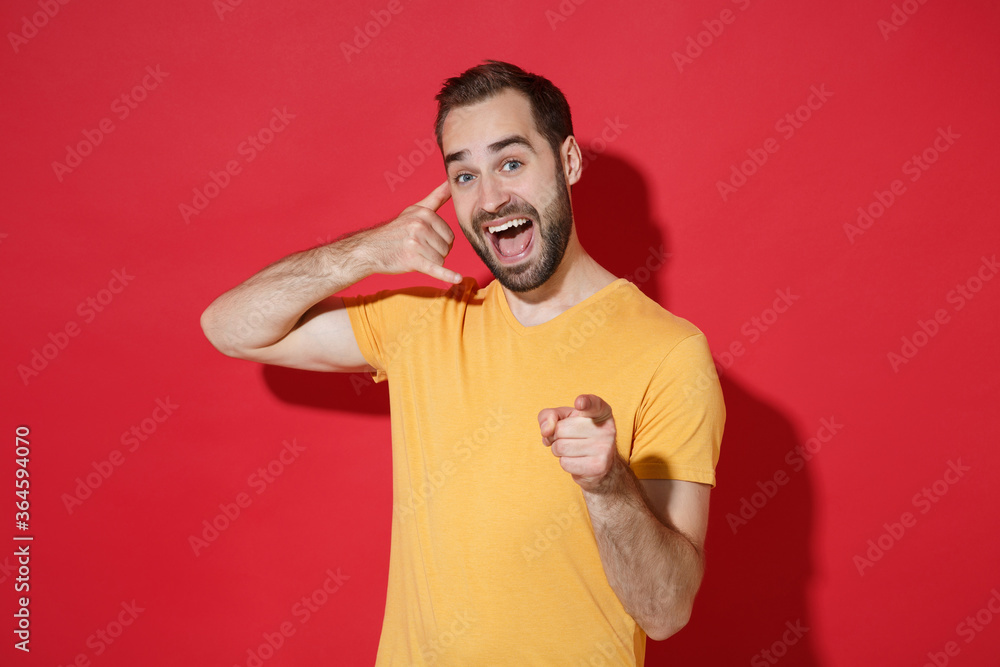 Excited young bearded guy in casual yellow t-shirt isolated on red background. People lifestyle concept. Mock up copy space. Doing phone gesture like says call me back pointing index finger on camera. Excited young bearded guy in casual yellow t-shirt isolated on red background. People lifestyle concept. Mock up copy space. Doing phone gesture like says call me back pointing index finger on camera.