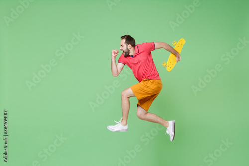full length Crazy young man guy in casual red pink t-shirt posing isolated on green background studio portrait. People lifestyle concept. Mock up copy space Jumping like running hold yellow skateboard