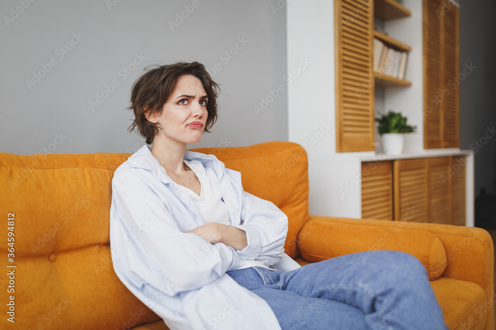 Puzzled boring young brunette woman in casual clothes sitting on couch spending time in living room at home. Rest relax good mood leisure lifestyle concept. Mock up copy space. Holding hands crossed. Puzzled boring young brunette woman in casual clothes sitting on couch spending time in living room at home. Rest relax good mood leisure lifestyle concept. Mock up copy space. Holding hands crossed.
