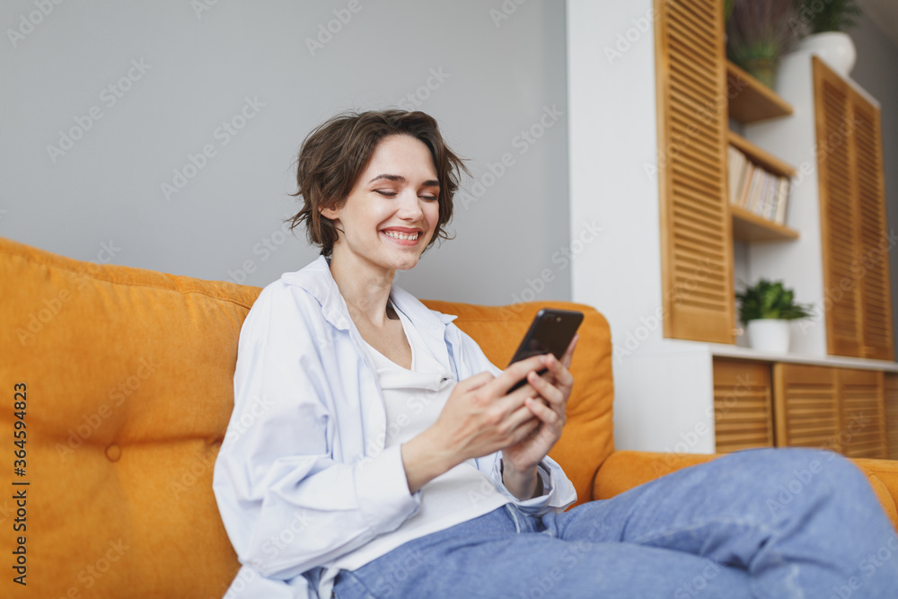 Smiling young woman girl in casual clothes sit on couch spending time in living room at home. Rest relax good mood leisure lifestyle concept. Mock up copy space. Using mobile phone typing sms message. Smiling young woman girl in casual clothes sit on couch spending time in living room at home. Rest relax good mood leisure lifestyle concept. Mock up copy space. Using mobile phone typing sms message.