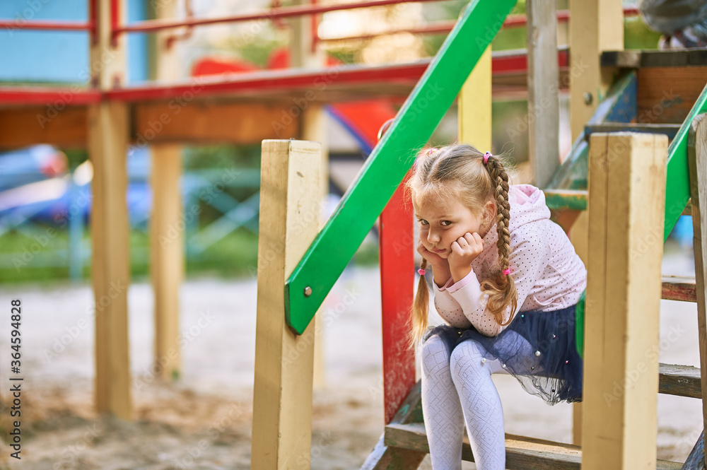 A little girl sits on the colored steps on the playground and thinks. Baby offended and sad in the playground A little girl sits on the colored steps on the playground and thinks. Baby offended and sad in the playground
