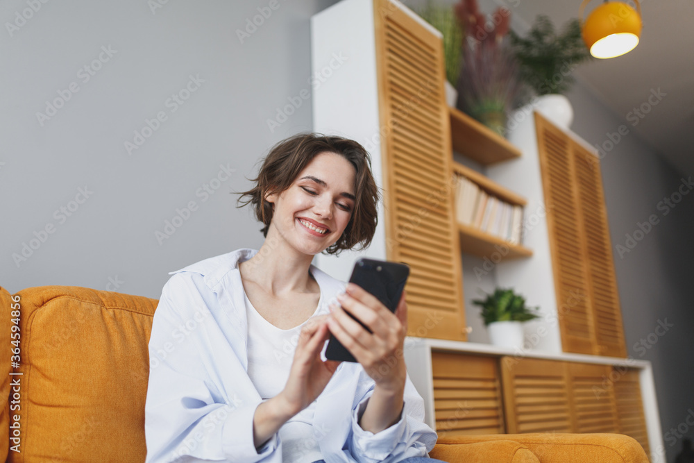 Smiling young woman girl in casual clothes sit on couch spending time in living room at home. Rest relax good mood leisure lifestyle concept. Mock up copy space. Using mobile phone typing sms message. Smiling young woman girl in casual clothes sit on couch spending time in living room at home. Rest relax good mood leisure lifestyle concept. Mock up copy space. Using mobile phone typing sms message.