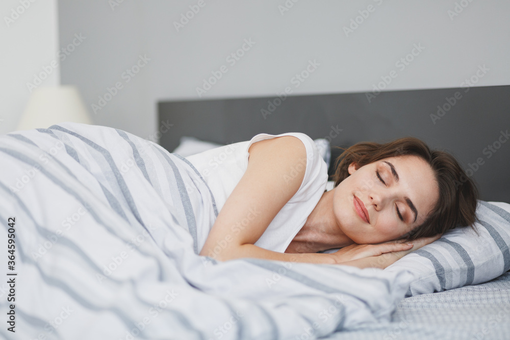 Beautiful attractive young woman in white t-shirt sleeping lying in bed with striped sheet pillow blanket spending time in bedroom at home. Rest relax good mood lifestyle concept. Mock up copy space. Beautiful attractive young woman in white t-shirt sleeping lying in bed with striped sheet pillow blanket spending time in bedroom at home. Rest relax good mood lifestyle concept. Mock up copy space.