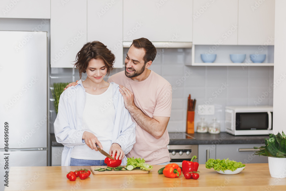 Smiling young couple two friends guy girl in casual clothes preparing vegetable salad cooking food in light kitchen at home. Dieting family healthy lifestyle concept. Mock up copy space. Hugging.