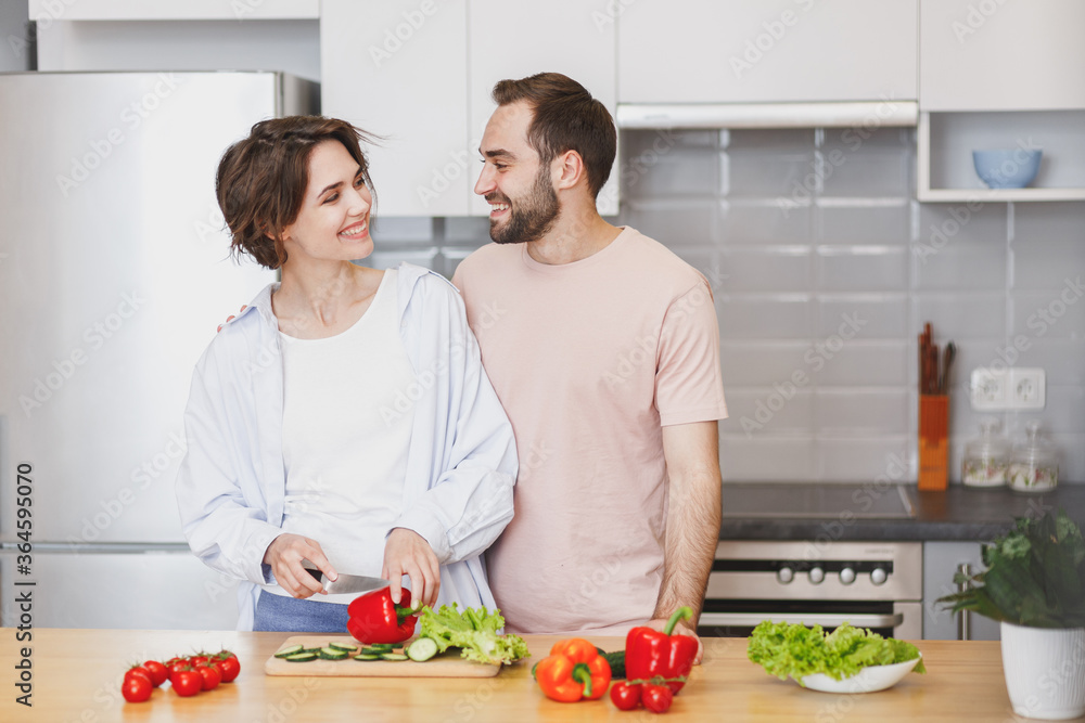 Smiling young couple friends guy girl in casual clothes preparing vegetable salad cooking food in kitchen at home. Dieting family healthy lifestyle concept. Mock up copy space. Looking at each other.