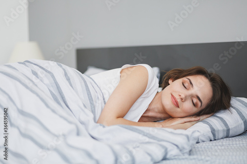 Beautiful attractive young woman in white t-shirt sleeping lying in bed with striped sheet pillow blanket spending time in bedroom at home. Rest relax good mood lifestyle concept. Mock up copy space.