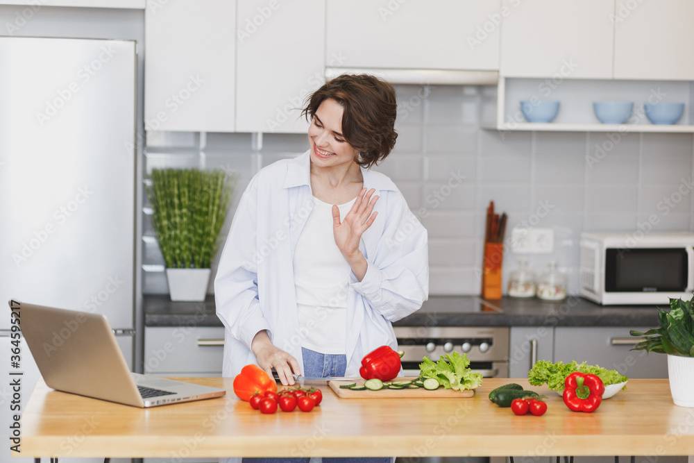 Smiling young housewife woman girl in white casual clothes using laptop computer making video call preparing vegetable salad cooking food in light kitchen at home. Dieting healthy lifestyle concept. Smiling young housewife woman girl in white casual clothes using laptop computer making video call preparing vegetable salad cooking food in light kitchen at home. Dieting healthy lifestyle concept.