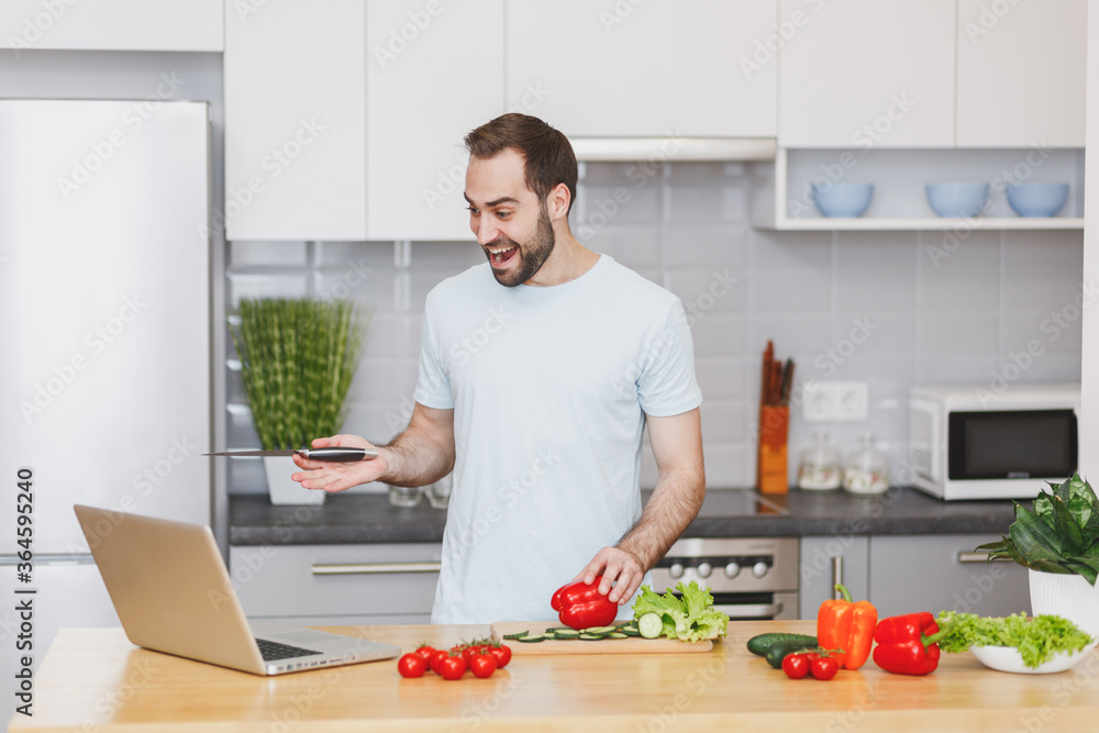 Excited young man guy in casual t-shirt using laptop computer making video call preparing vegetable salad cooking food in light kitchen at home. Dieting healthy lifestyle concept. Mock up copy space. Excited young man guy in casual t-shirt using laptop computer making video call preparing vegetable salad cooking food in light kitchen at home. Dieting healthy lifestyle concept. Mock up copy space.