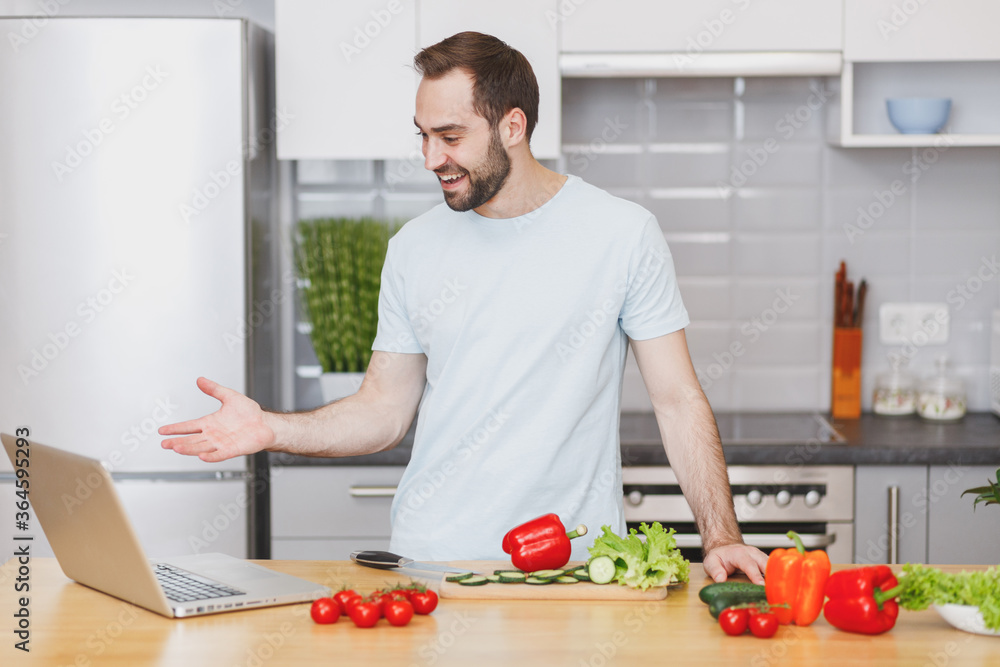 Smiling young man guy in casual t-shirt using laptop computer making video call preparing vegetable salad cooking food in light kitchen at home. Dieting healthy lifestyle concept. Mock up copy space. Smiling young man guy in casual t-shirt using laptop computer making video call preparing vegetable salad cooking food in light kitchen at home. Dieting healthy lifestyle concept. Mock up copy space.