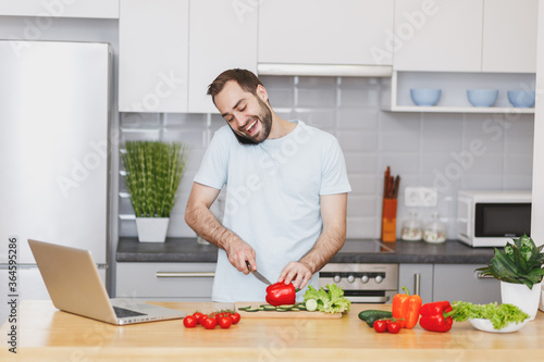 Cheerful young bearded man guy in white casual t-shirt talking on mobile phone preparing vegetable salad cooking food in light kitchen at home. Dieting healthy lifestyle concept. Mock up copy space.