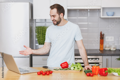 Smiling young man guy in casual t-shirt using laptop computer making video call preparing vegetable salad cooking food in light kitchen at home. Dieting healthy lifestyle concept. Mock up copy space.