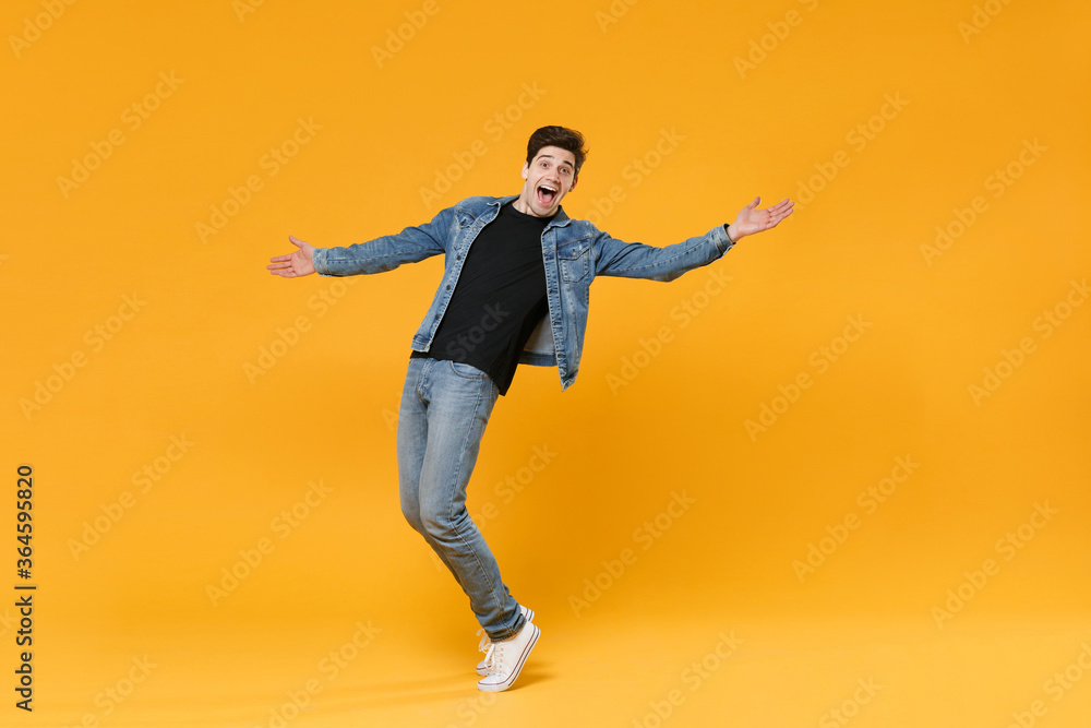full length Excited young man guy in casual denim clothes posing isolated on yellow wall background studio portrait. People emotions lifestyle concept. Mock up copy space Stand on toes spreading hands