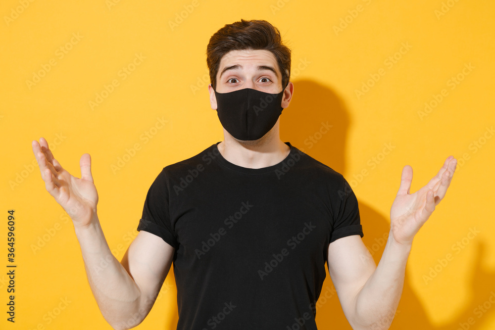 Shocked young man guy in black t-shirt face mask isolated on yellow wall background studio portrait. Epidemic pandemic spreading coronavirus 2019-ncov sars covid-19 flu virus concept. Spreading hands. Shocked young man guy in black t-shirt face mask isolated on yellow wall background studio portrait. Epidemic pandemic spreading coronavirus 2019-ncov sars covid-19 flu virus concept. Spreading hands.