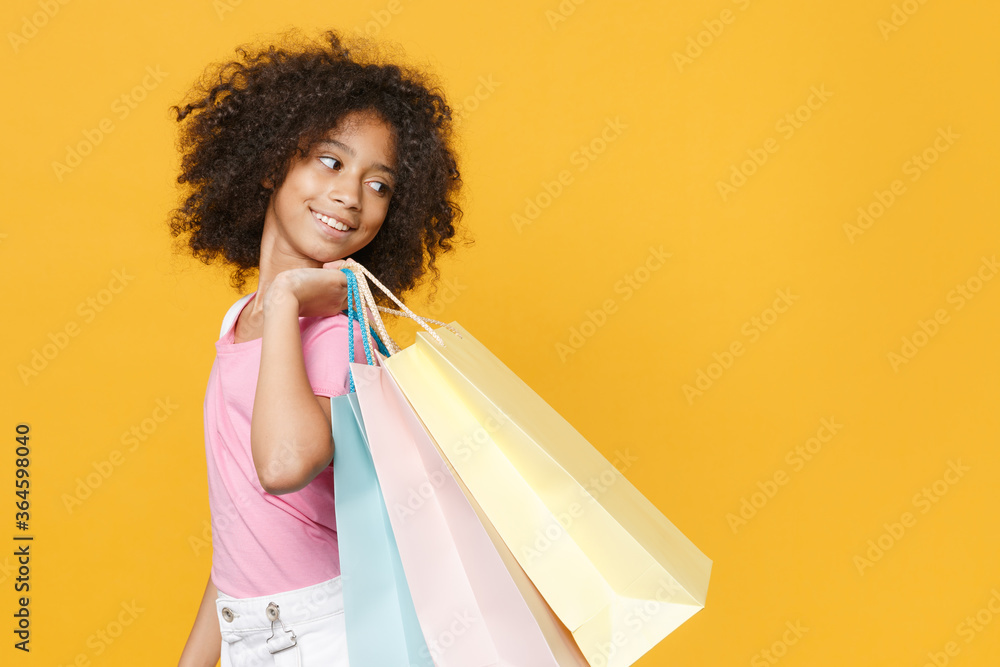 Side view of pretty little african american kid girl 12-13 years old in pink t-shirt isolated on yellow wall background. Childhood lifestyle concept. Hold package bag with purchases after shopping. Side view of pretty little african american kid girl 12-13 years old in pink t-shirt isolated on yellow wall background. Childhood lifestyle concept. Hold package bag with purchases after shopping.
