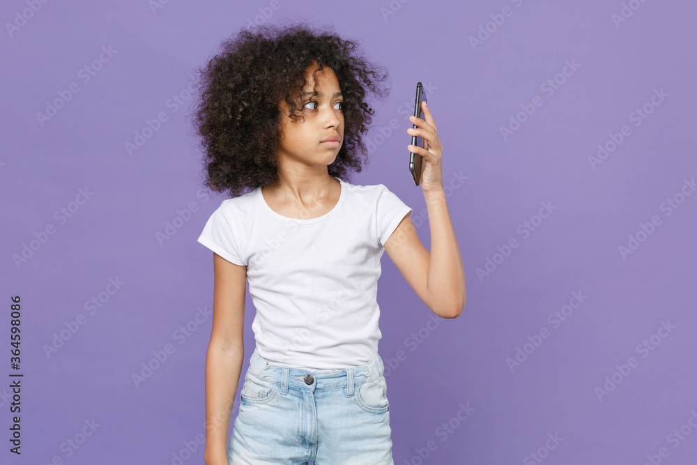 Perplexed little african american kid girl 12-13 years old in white t-shirt isolated on violet background studio portrait. Childhood lifestyle concept. Mock up copy space. Talking on mobile phone. Perplexed little african american kid girl 12-13 years old in white t-shirt isolated on violet background studio portrait. Childhood lifestyle concept. Mock up copy space. Talking on mobile phone.