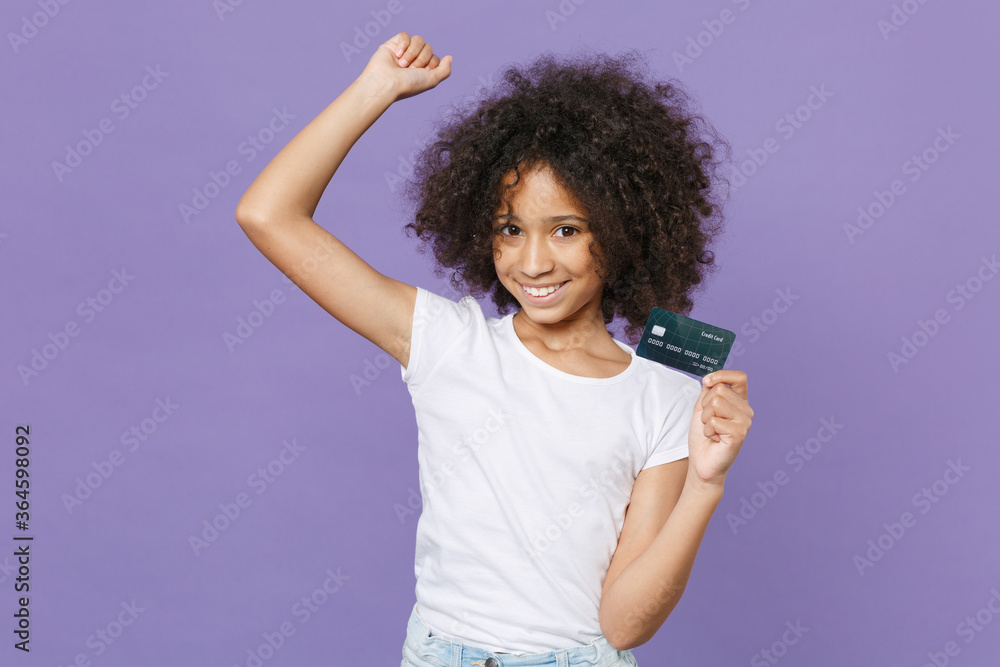 Joyful little african american kid girl 12-13 years old in white t-shirt isolated on violet background. Childhood lifestyle concept. Mock up copy space. Hold credit bank card, doing winner gesture.