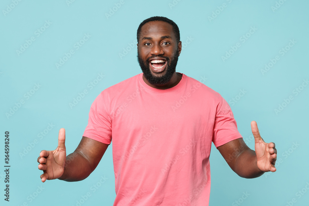 Fototapeta premium Excited young african american man guy in casual pink t-shirt isolated on blue background studio. People lifestyle concept. Mock up copy space. Gesturing demonstrating size with horizontal workspace.