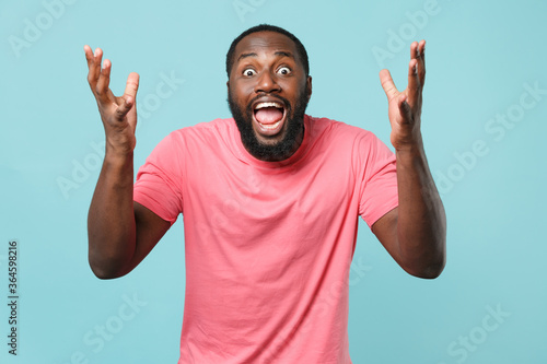 Shocked worried young african american man guy in casual pink t-shirt isolated on blue background studio portrait. People emotions lifestyle concept. Mock up copy space. Spreading hands screaming.