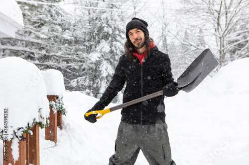 young caucasian man with a smile, standing outdoor holding a shovel to shoveling the snow. house and trees covered by snow in the background.