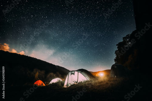 Summer camping in the mountains. Tents in the night with the starry sky and clouds in the background. Illuminated tents on the grassy ground in the mountains.